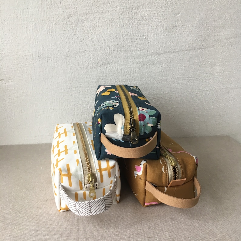 May include: Three zippered pouches with fabric handles. The pouch on top has a floral pattern in shades of blue, green, and white. The middle pouch has a yellow and white geometric pattern. The bottom pouch has a brown and white geometric pattern.