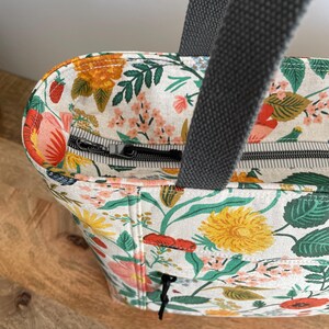 May include: A white tote bag with a floral print featuring red, yellow, and green flowers. The bag has a zipper closure and two gray canvas straps.