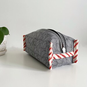 May include: A black and white patterned makeup bag with red and white striped trim. The bag has a zipper closure.