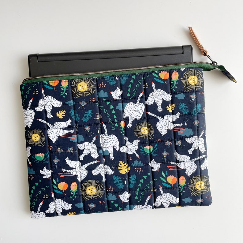 Puede incluir: Una funda para port&aacute;til de color azul oscuro con un dise&ntilde;o acolchado que presenta cisnes blancos y patrones florales coloridos. La funda tiene una cremallera y una leng&uuml;eta de cuero marr&oacute;n.