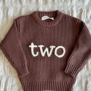 May include: A brown knit sweater with the word "two" in white, block letters across the front. The sweater has a ribbed texture and a crew neck. The sweater is likely for a toddler.
