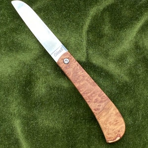 May include: A small, silver pocket knife with a wooden handle. The handle is a light brown color with a dark brown marbled pattern. The blade is partially open and the knife is on a green fabric surface. The knife has the text "Giless" engraved on the blade.