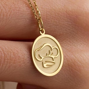 May include: A gold-colored pendant necklace featuring an oval charm with a chef's hat design. The pendant hangs from a delicate chain. The chef's hat is embossed within the oval frame.
