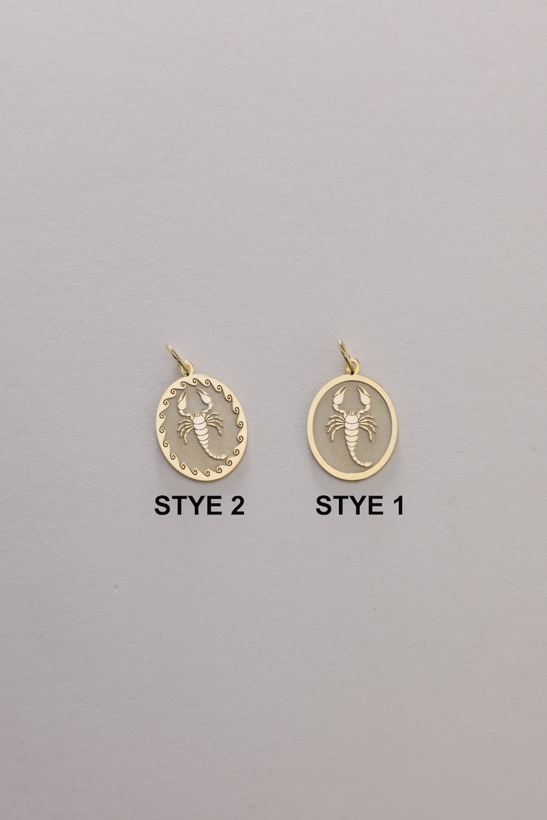 May include: Two gold-toned pendants featuring a scorpion design. The pendant on the left has a more intricate border, while the pendant on the right has a simpler border. The text "STYE 2" is below the left pendant, and the text "STYE 1" is below the right pendant.
