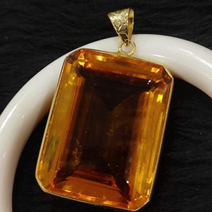 May include: A rectangular pendant with a large, faceted, amber-colored gemstone set in a gold-colored frame. The pendant has a decorative gold bail. The background is dark, with a white curved object partially visible.