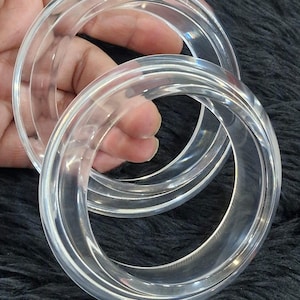 May include: Two clear, transparent acrylic bangles. The circular bracelets are stacked, showcasing their smooth, polished surfaces. The bangles are held against a dark, textured background.