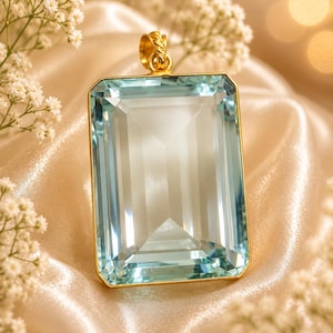 Peut inclure: Pendentif rectangulaire en or avec une grande pierre précieuse bleu clair. La pierre est taillée à facettes et sertie dans un cadre doré. Le pendentif est suspendu à une bélière en or. Fond satiné avec des fleurs blanches.