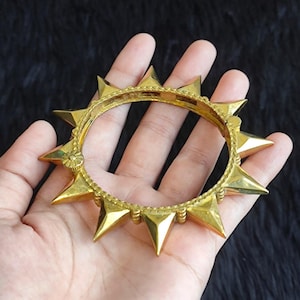 Puede incluir: Un brazalete circular de tono dorado que presenta una serie de puntas triangulares prominentes que se extienden hacia afuera. La banda tiene un borde texturizado con cuentas e incluye un pequeño elemento decorativo circular en un lado.