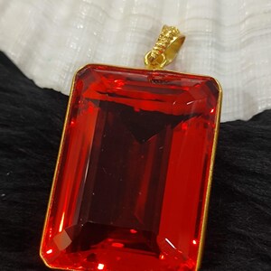 May include: A rectangular, faceted red gemstone pendant with a gold-colored frame and bail. The pendant is a deep, vibrant red color and appears to be made of glass or a similar material. The bail has a textured design.
