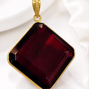 May include: A gold-toned pendant with a large, square, deep red gemstone. The gemstone is set in a gold frame and has a faceted appearance. The pendant has a textured bail for a chain.