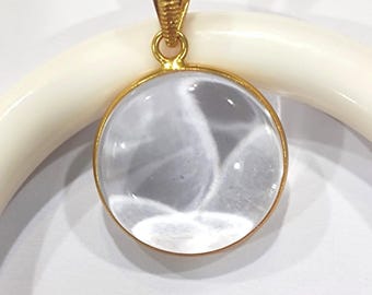 May include: A clear, round pendant with a gold-colored bezel and bail. The pendant has a gold-colored loop at the top for a chain. The pendant is set against a white background.