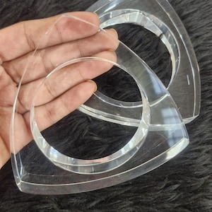 May include: Two clear acrylic purse handles with a triangular shape and a circular opening. The handles are transparent and appear to be made of a thick, sturdy material. The handles are shown against a dark, textured background.