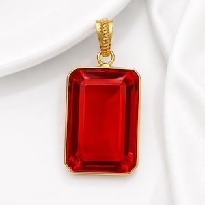 May include: A rectangular red pendant with a gold-colored frame and bail. The pendant has a large, faceted, ruby-like appearance. The bail is textured with a rope-like design. The background is white.