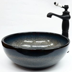 May include: A black ceramic vessel sink with a speckled blue and white glaze. The sink is round and has a wide, shallow bowl. A black faucet with a white ceramic handle is mounted above the sink.