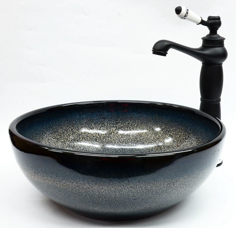 May include: A black ceramic vessel sink with a speckled blue and white glaze. The sink is round and has a wide, shallow bowl. A black faucet with a white ceramic handle is mounted above the sink.