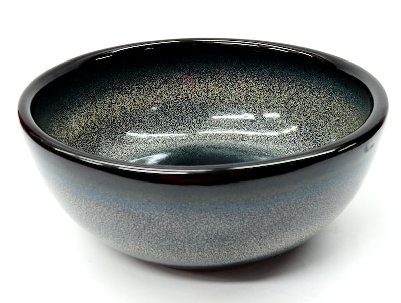 May include: A black ceramic bowl with a speckled, blue and gray glaze. The bowl has a rounded shape and a wide rim.