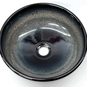 May include: A black ceramic vessel sink with a white speckled glaze. The sink has a round shape and a single drain hole in the center.