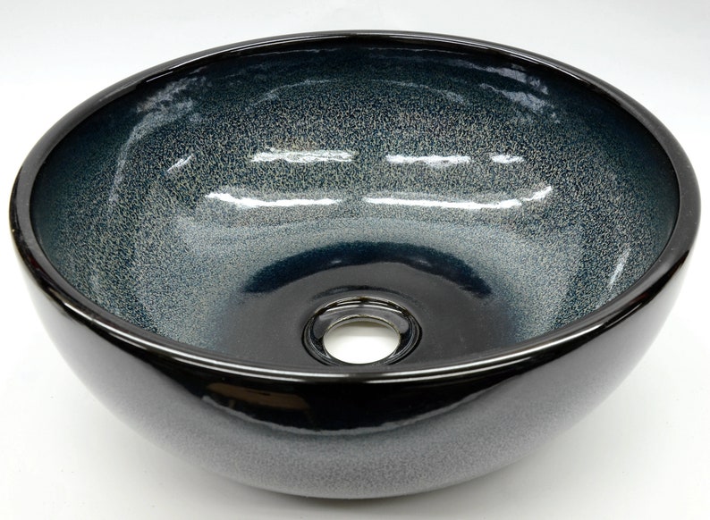 May include: A round, black ceramic vessel sink with a speckled, blue glaze. The sink has a single drain hole in the center.