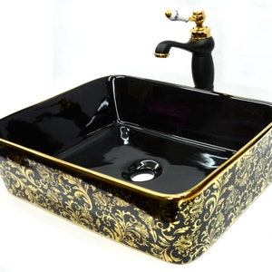 May include: Black ceramic vessel sink with a gold rim and a gold floral pattern. The sink is rectangular and has a single drain hole. A black faucet with a gold handle is mounted above the sink.