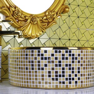 May include: A gold mosaic-tiled bathroom sink with a matching gold faucet and a decorative gold-framed mirror. The sink is cylindrical and features a pattern of gold and white tiles. The faucet has a white and gold handle.