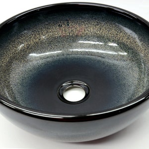 May include: A round, black ceramic vessel sink with a subtle gradient of gray and gold. The sink has a single drain hole in the center.