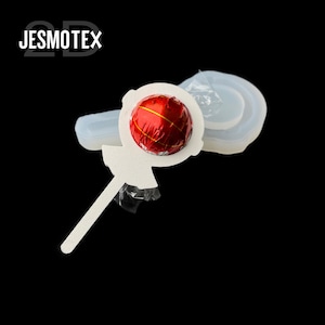May include: A white lollipop-shaped mould with a red and gold wrapped sweet. The sweet is in the centre of the mould, which has a handle. The word "JESMOTEX" is in the upper left corner of the image.