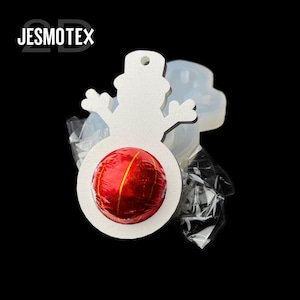 May include: A white snowman-shaped decoration with a red and gold wrapped sweet in the centre. The decoration has a hole at the top for hanging. The image also shows a clear mould and the text "JESMOTEX".