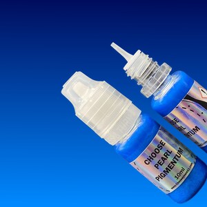 May include: Two small plastic squeeze bottles filled with vibrant blue pearl pigment. Each bottle features a clear cap and a label with the text CHOOSE PEARL PIGMENTUM 10ml. The bottles are positioned against a solid blue background.