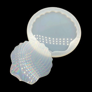 May include: A clear, seashell-shaped silicone mold with a scalloped edge. The mold is designed for crafting and features a detailed seashell design. The image shows the mold and a cast seashell, both with iridescent highlights, against a black background.