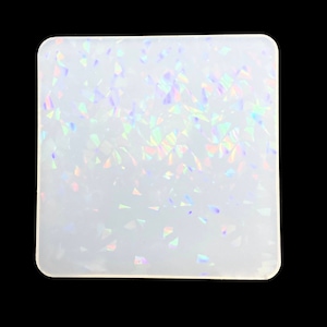 Puede incluir: Un posavasos cuadrado blanco con esquinas redondeadas. La superficie está cubierta de pequeñas formas reflectantes de colores que brillan con un efecto arcoíris. El posavasos está sobre un fondo negro.