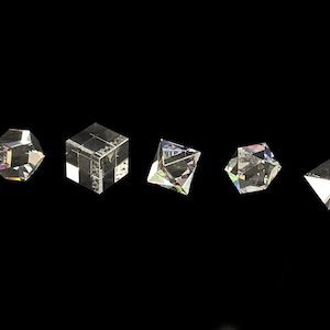 Può includere: Cinque prismi geometrici in vetro trasparente disposti in una fila orizzontale su uno sfondo nero. Le forme includono una sfera sfaccettata, un cubo, una piramide, un prisma esagonale e una piramide triangolare che rifrangono la luce.
