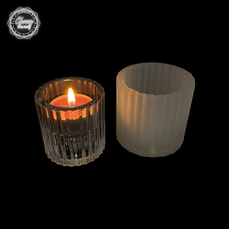 May include: A lit votive candle in a clear, ribbed glass votive holder next to an empty, unlit votive holder. The candle flame glows brightly against the dark background. The votive holders are cylindrical.