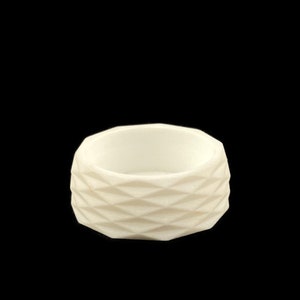 May include: A small, off-white, geometric-patterned container. The container has a cylindrical shape with a wide opening and a textured exterior featuring a repeating diamond pattern. The background is solid black.