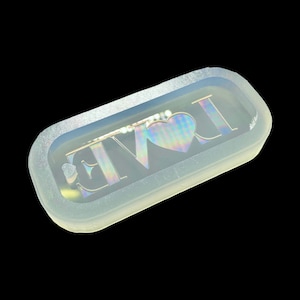 May include: A clear silicone mould for resin casting. The mould is shaped like a rectangle with rounded corners and features the word "LOVE" with a heart in the centre.