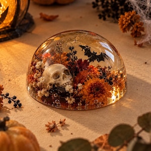 Könnte beinhalten: Ein klarer, kuppelförmiger Briefbeschwerer aus Kunstharz, der einen Miniatur-Totenkopf, eine schwarze Fledermaussilhouette und konservierte herbstfarbene Blumen wie orangefarbene Chrysanthemen und weißes Schleierkraut in sich einschließt.