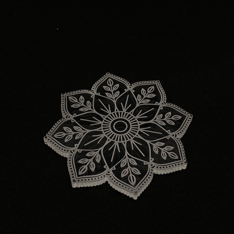 May include: A white, laser-cut mandala design with intricate details, resembling a flower with multiple petals and leaves.