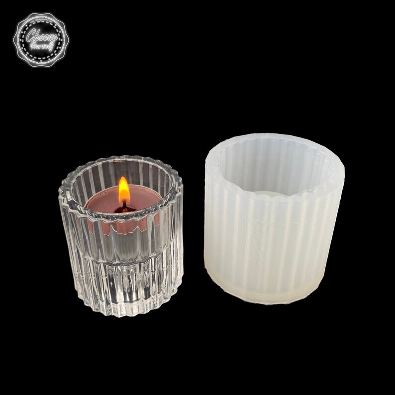 May include: A clear glass votive candle holder with a lit candle and a white silicone mold. The candle holder has vertical ribbed detailing. The candle has a small flame. The silicone mold is cylindrical with vertical ribbed detailing.