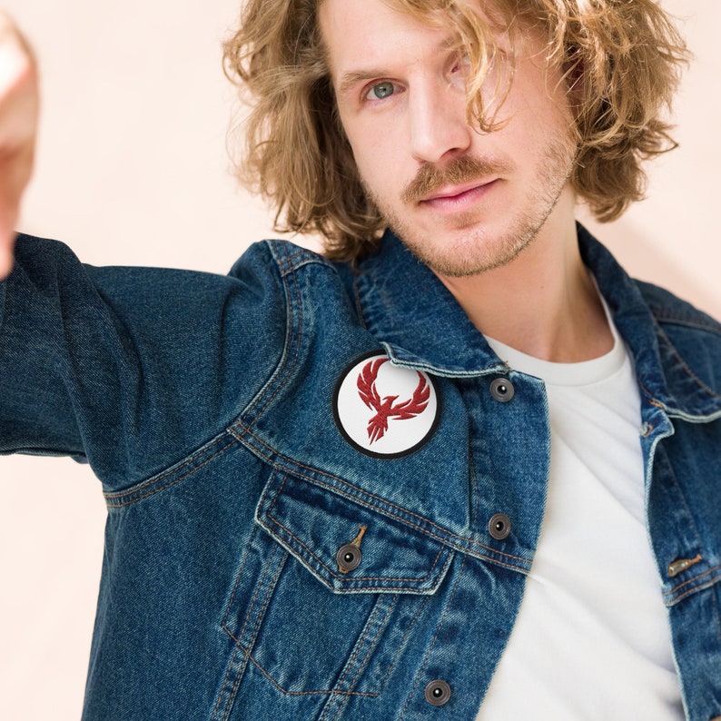 May include: A round patch featuring a red phoenix design on a white background, with a black border. The patch is affixed to a blue denim jacket.