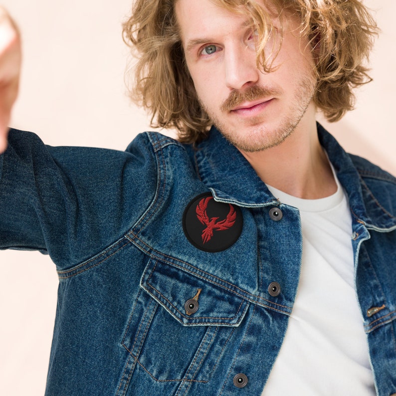 May include: A dark blue denim jacket with a black circular patch featuring a red phoenix design. The jacket has a classic design with button closures and pockets. The patch is embroidered with a detailed bird motif.