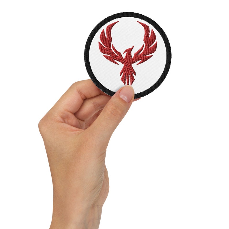 May include: Round embroidered patch featuring a red phoenix design on a white background, framed by a black border. The phoenix is depicted with outstretched wings and a stylized form. This patch is suitable for clothing or accessories.