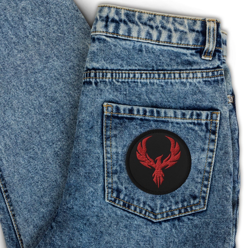May include: A pair of light blue denim jeans with a black circular patch on the back pocket. The patch features a red phoenix design embroidered on a black background. The jeans have a classic five-pocket design.
