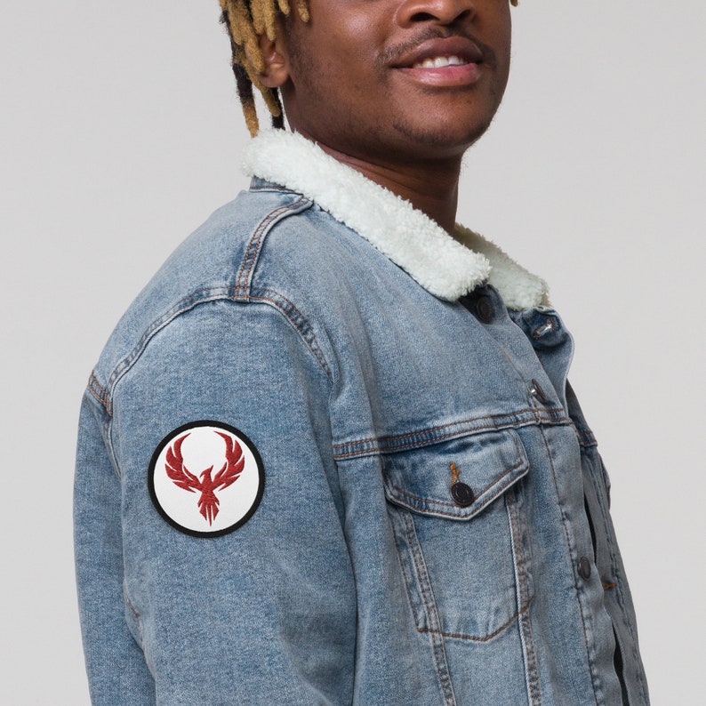 May include: A light blue denim jacket with a white sherpa collar. A circular patch with a red phoenix design is on the left sleeve. The jacket has two chest pockets and a button closure.