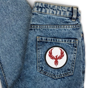 May include: A round patch featuring a red phoenix design on a white background, with a black border. The patch is sewn onto the back pocket of a pair of light blue denim jeans.