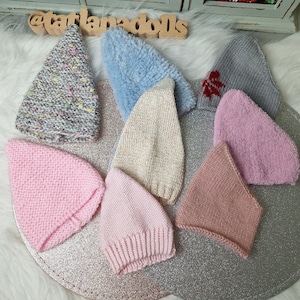 May include: Assortment of 10 knitted hats in various colors and textures, including pink, blue, beige, and gray. The hats are arranged on a white surface with a wooden sign that reads "@tatlanadolls".