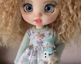 May include: A doll with large blue eyes and blonde curly hair. The doll wears a dress with a deer pattern and a small snowman. The dress has a light blue and white floral pattern. The doll is holding a small snowman.