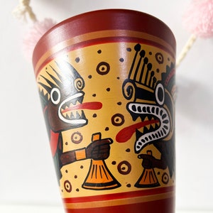 Pode incluir: Uma caneca de cerâmica vermelha com um desenho amarelo, laranja, preto e branco de duas criaturas míticas com a boca aberta e sinos nas mãos.