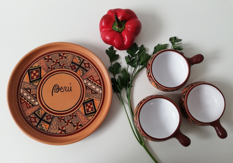 May include: A set of three small, white ceramic bowls with brown handles and geometric designs. The bowls are arranged on a white surface with a larger, brown ceramic plate with a geometric design and the word "Per&uacute;" written in the center. A red bell pepper and a sprig of parsley are also on the surface.