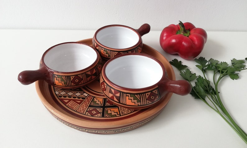 May include: Set of three small ceramic bowls with handles, decorated with geometric patterns in brown, orange, and green. The bowls are arranged on a larger ceramic plate with a similar design. A red bell pepper and a bunch of parsley are in the background.