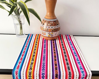 Op de afbeelding: Een kleurrijke tafelloper met verticale strepen in verschillende kleuren, waaronder rood, blauw, geel en paars. Een bruine vaas met geometrische patronen staat erop. Links staat een transparante vaas met groene planten.
