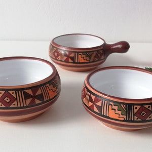 May include: Three small terracotta bowls with white interiors and brown handles. The bowls are decorated with geometric patterns in orange, brown, green, and red.